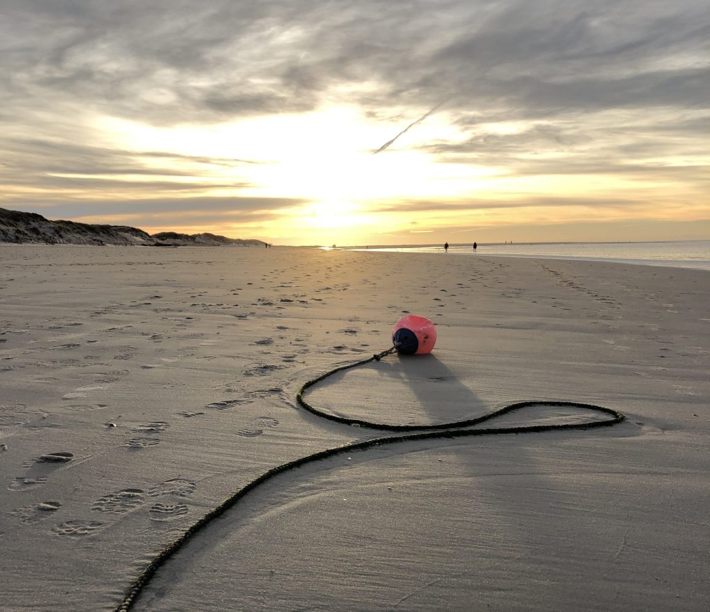 Ein langer, geschwungener Ankerseilstrang mit roter Boje im Sand eines weiten, menschenleeren Strandes bei Sonnenuntergang, Symbol für Ankommen und innere Ruhe.