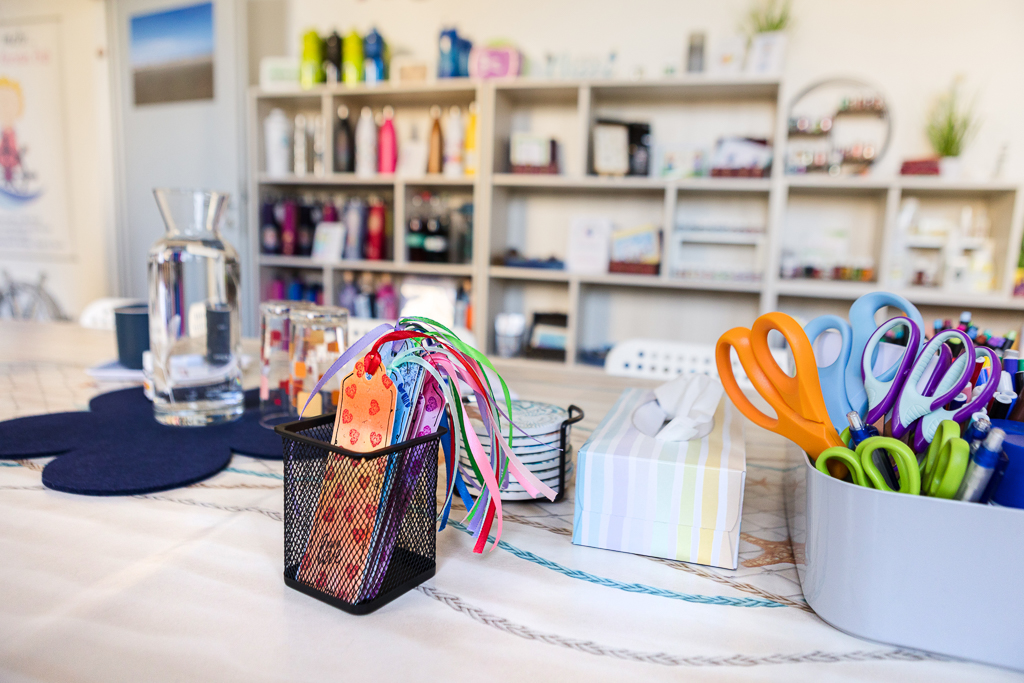 Nahaufnahme eines Kreativtisches mit Scheren, bunten Bändern, Papieranhängern, Wasserkrug und Bastelmaterial im Studio „Räume für mehr“ in Nürnberg. Im Hintergrund Regale mit Produkten und Pflanzen.