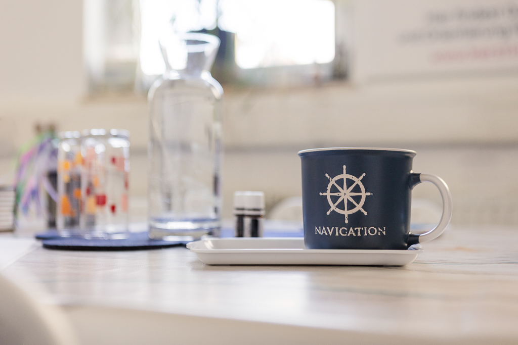 Nahaufnahme einer dunkelblauen Tasse mit Steuerrad und dem Schriftzug „Navigation“ auf einem Tisch. Daneben Glas und kleine Fläschchen. Symbol für Orientierung und Wegbegleitung im Studio „Räume für mehr…“ in Nürnberg.