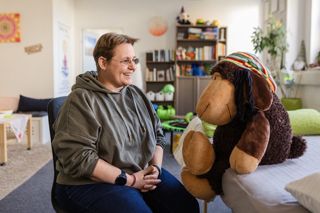 Gespräche auf Augenhöhe – für Kinder, Jugendliche und Erwachsene Bild: Nina Thilo | Auf dem Bild sieht man eine freundliche, einladende Szene in einem gemütlich eingerichteten Raum. Eine Frau sitzt lächelnd einem großen Stofftier gegenüber.