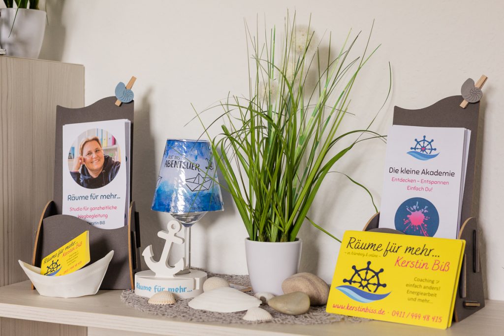 Flyer zu Räume für mehr und Die kleine Akademie im kleinen Kauflädchen, dekoriert mit einer handbemalten Lampe, Muscheln und Pflanzen.