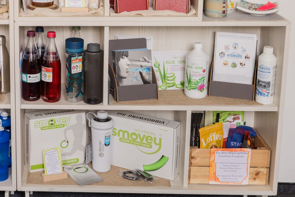 Regal im kleinen Kauflädchen mit smovey®-Ringen, Blender Bottle, Q-Life Aloe Vera-Produkten und Hydroxil. Rechts unten eine Kiste mit reduzierten Einzelstücken – Schnäppchen aus dem Regal gefallen.